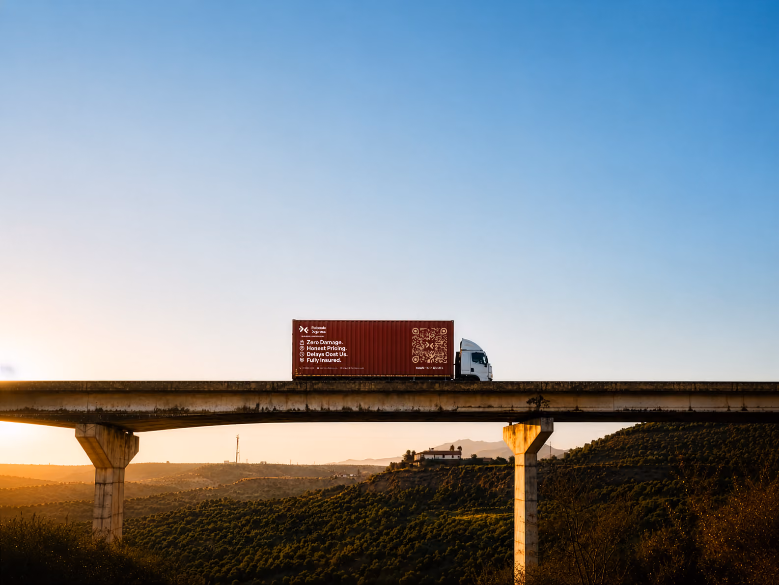 Semi-truck with cargo crossing elevated highway bridge at sunset over desert landscape